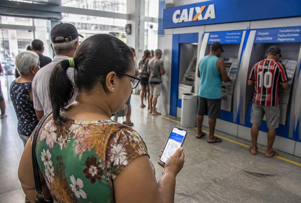 Beneficiária consultando o aplicativo do banco em agência da Caixa Econômica Federal enquanto outras pessoas usam caixas eletrônicos